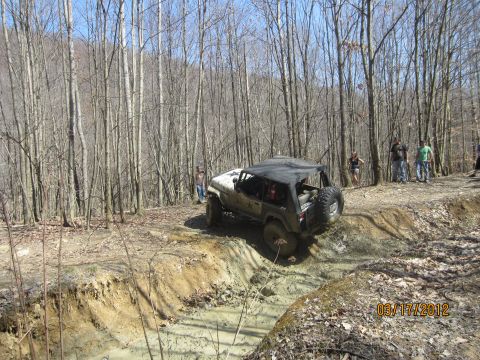 1979 Ford Bronco - A Jeep Wrangler tackles a challenging muddy off-road course surrounded by trees.
