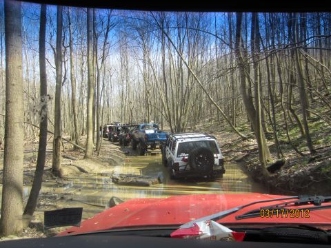 1979 Ford Bronco - A convoy of off-road vehicles drives through a muddy trail in a wooded area.