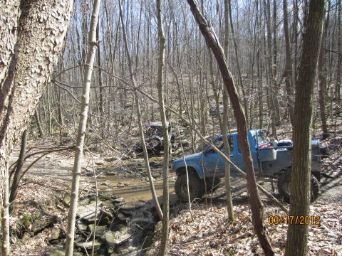 1979 Ford Bronco - Two off-road vehicles navigate a muddy path through a dense forest with bare trees.