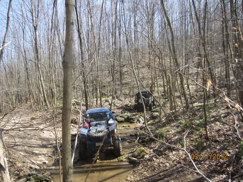 Two off-road vehicles navigating a rocky stream bed in a wooded area