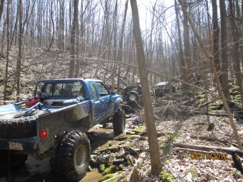 1979 Ford Bronco - Multiple off-road vehicles traverse a challenging wooded terrain with water and rocks.