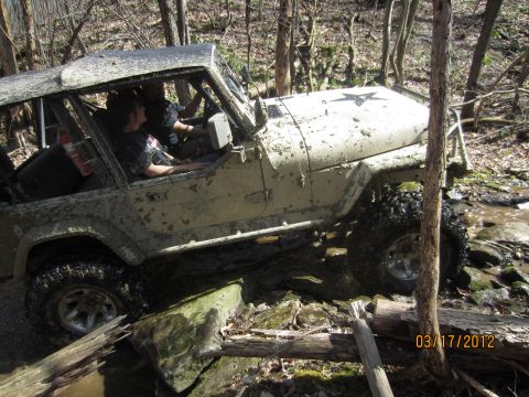 1979 Ford Bronco - A heavily muddied Jeep CJ-7 tackles a challenging off-road course with a driver and passenger inside.