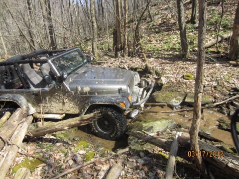 1979 Ford Bronco - A gray Jeep Wrangler is shown navigating a challenging off-road course through a wooded area with a muddy creek and fallen logs.