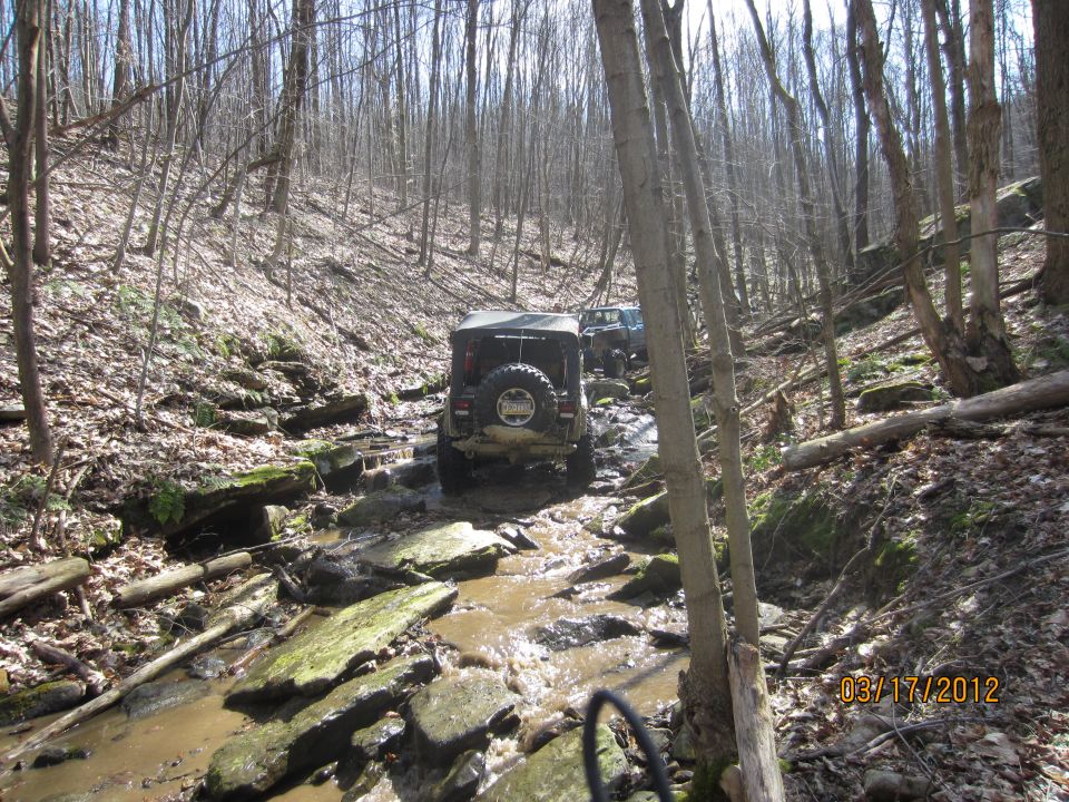 Two Jeeps navigating a muddy, rocky stream in a wooded area. Date stamp reads 03/17/2012