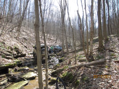 1979 Ford Bronco - Off-road vehicles navigate a challenging path through a wooded creek bed with rocky terrain.