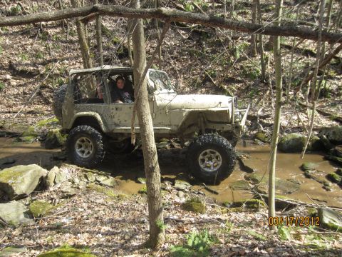 1979 Ford Bronco - A mud-covered Jeep Wrangler navigates a rocky creek bed surrounded by trees.