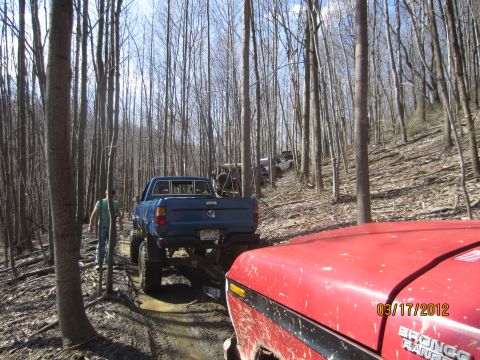 1979 Ford Bronco - Multiple off-road vehicles navigate a muddy trail through a dense forest on a sunny day.