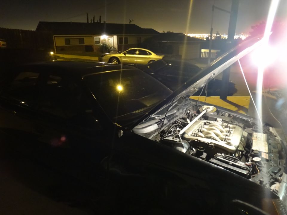 1993 Ford Taurus engine bay with hood open at night. Another car is parked in the background