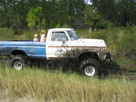1991 Ford Bronco - A white and blue pickup truck is stuck in the mud with two children in the back.