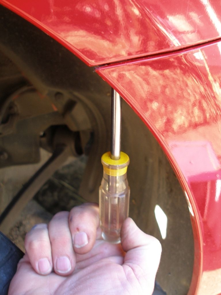 Hand using screwdriver on red car's fender liner near wheel well. Supermotors.net logo visible