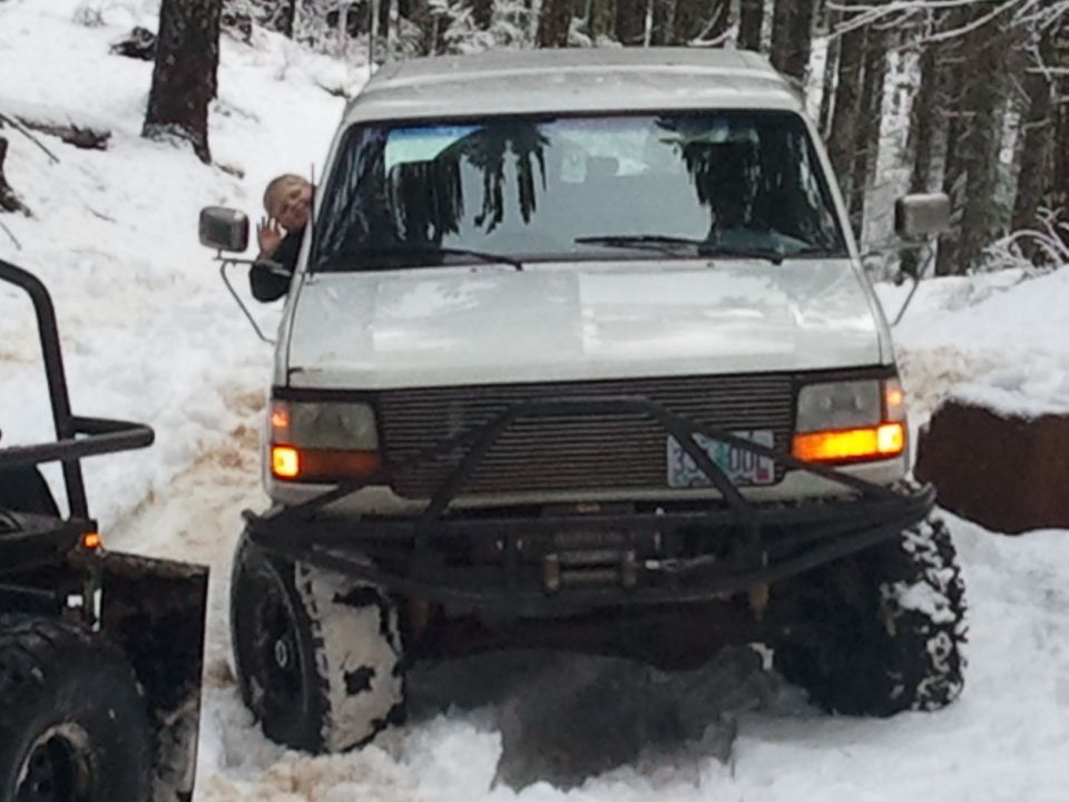 White van with custom bumper and winch in snow, person waving from window. Another vehicle partially visible on left