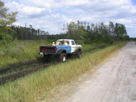 1991 Ford Bronco - Ford pickup truck with passengers in the bed navigates a muddy off-road path.