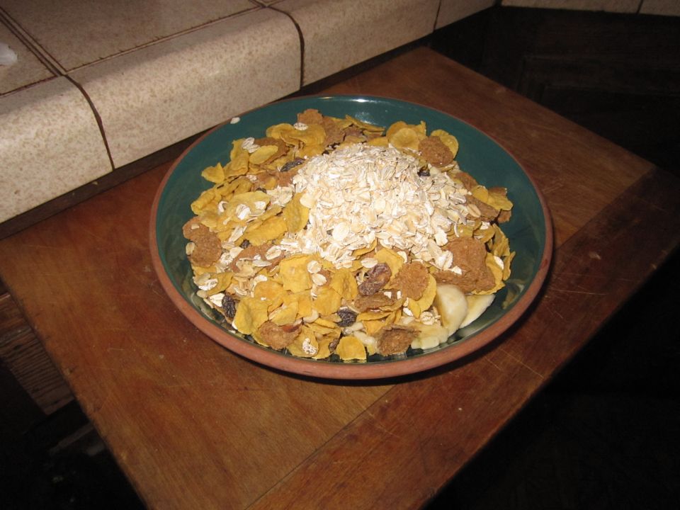 Bowl of cereal containing corn flakes, raisins, banana slices, and oats on a wooden table