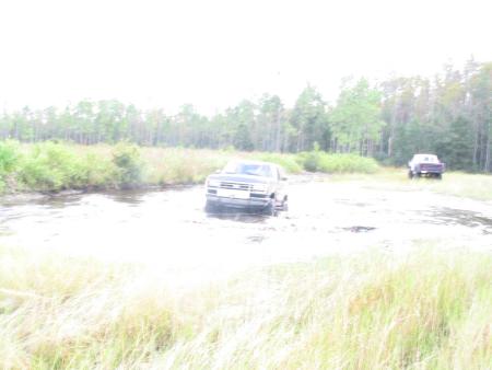 1991 Ford Bronco - Two trucks navigate through a watery landscape, with one vehicle in the foreground and another further back.