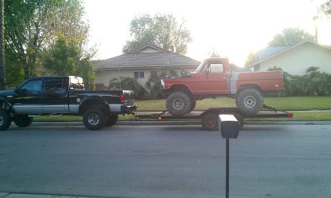 1969 Ford F-250 - A black Ford pickup truck tows a red Ford pickup truck on a trailer down a suburban street.