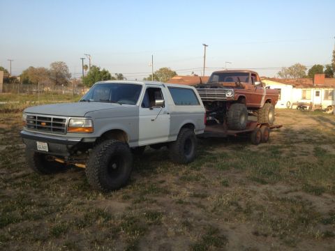 1969 Ford F-250 - A lifted Ford Bronco tows a vintage Ford F-250 on a trailer under a clear sky.