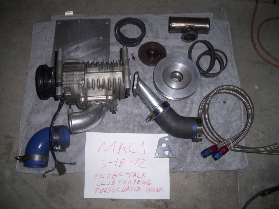 Supercharger assembly with hoses, belts, and mounting plate. Paper note reads 'MAC1 3-18-12 PROBE TALK CLUB PROTEGE PERFORMANCE PROBE'