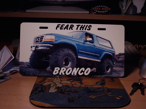 1996 Ford Bronco - A custom license plate a lifted blue Bronco with large tires and text is displayed on a desk.