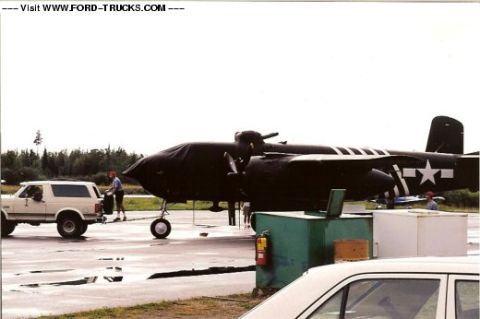 1992 Ford Bronco - A white SUV is parked on an airfield next to a large, tarp-covered aircraft.