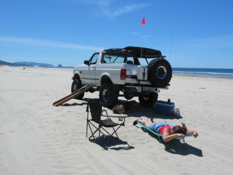 1995 Ford Bronco - A white 1995 Ford Bronco is parked on the sand near the ocean, with a person relaxing on a lounge chair.