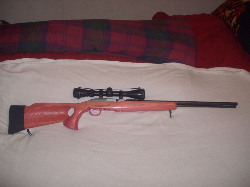 Rifle with pink laminated thumbhole stock and black scope lying on a white blanket on a bed