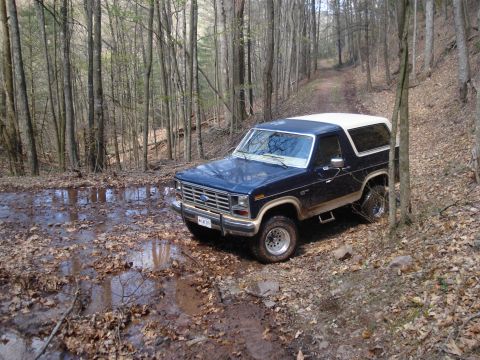 1985 Ford Bronco - A dark blue and white 1985 Ford Bronco navigates a muddy, wooded trail.
