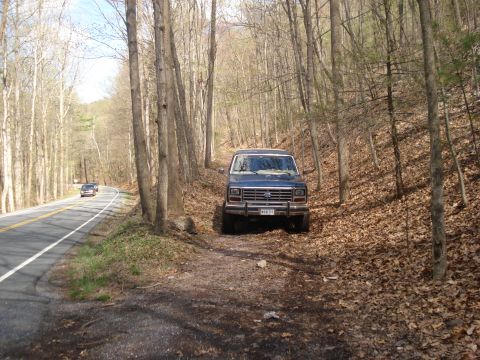 1985 Ford Bronco - A blue Ford Bronco is parked on a dirt path beside a winding road through a wooded area.