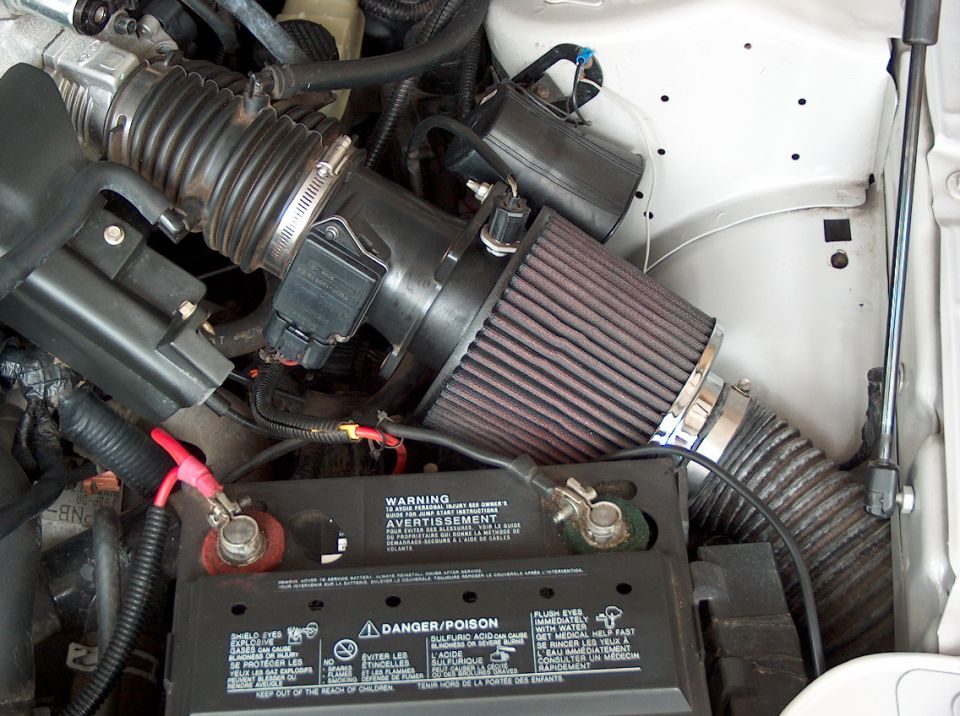 Engine bay with K&N cone air filter on throttle body, red top-post battery below with warning label
