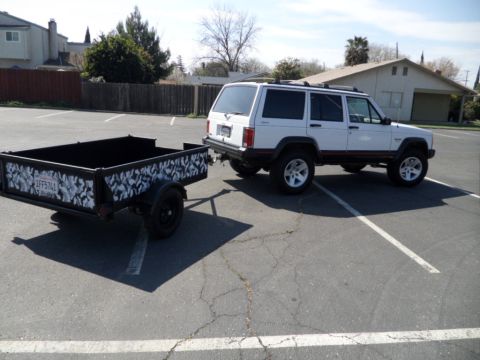 1994 Jeep Cherokee - A white Jeep Cherokee is parked in a lot, attached to a black utility trailer with a unique design.