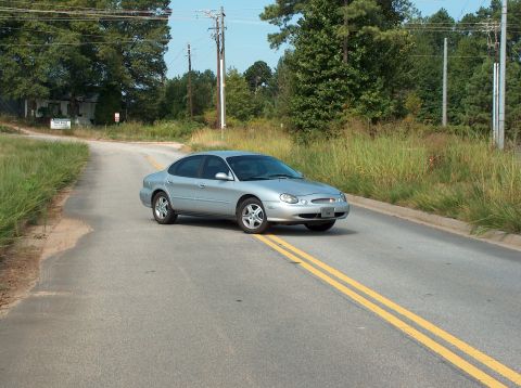 1998 Ford Taurus - A silver sedan drives down a rural road, its rounded body style suggesting a model year around 2000.