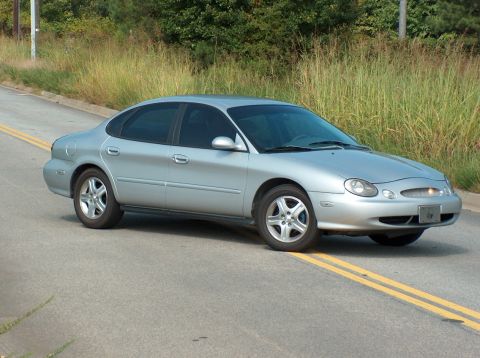 1998 Ford Taurus - This silver 1998 Ford Taurus sedan is captured from a three-quarter front view on a paved road with yellow lane markings.
