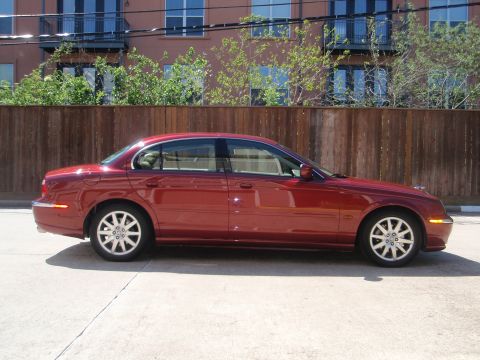 2000 Jaguar S-Type - A red 2000 Jaguar S-Type sedan is viewed from its side, parked in front of a wooden fence and a building.