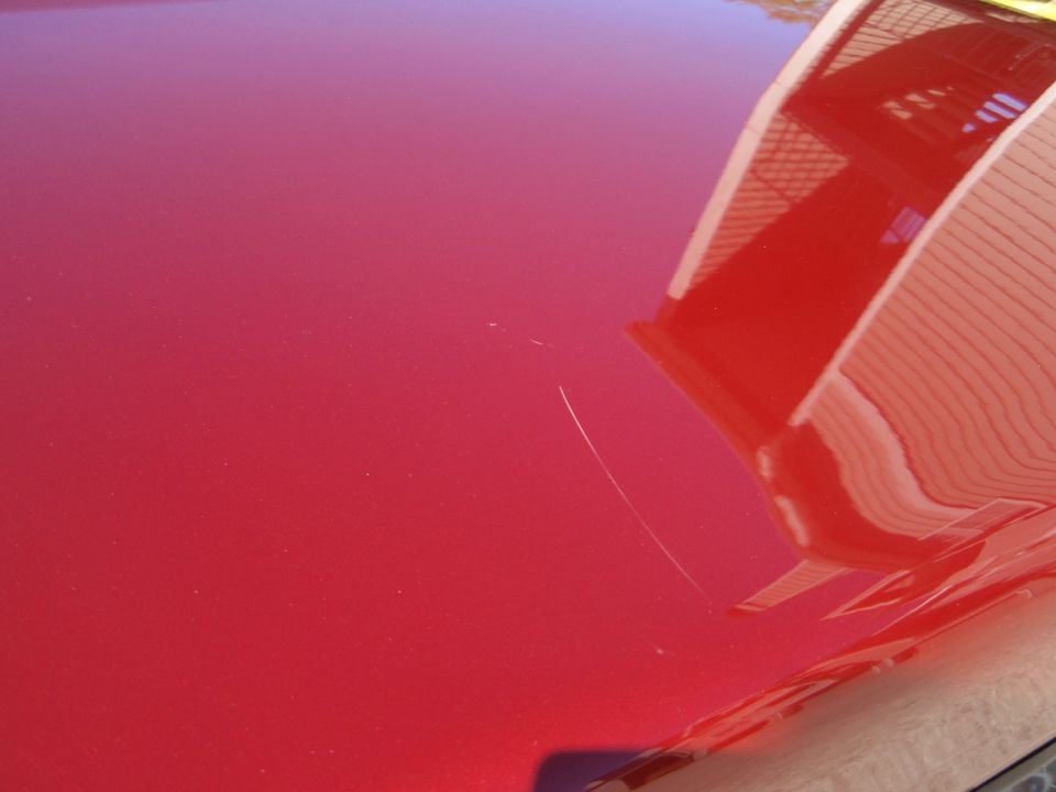 Red car hood with a prominent scratch and reflection of a building