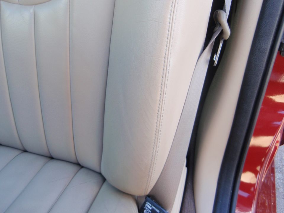Beige leather car seat with stitching and a gray seatbelt. Red exterior visible on the right