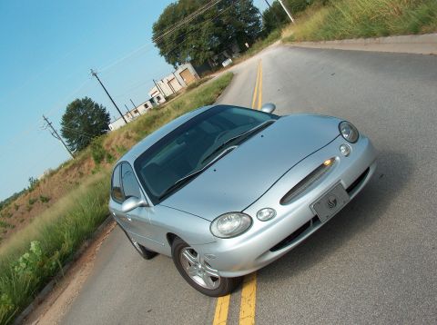 1998 Ford Taurus - This silver sedan is parked on the side of a road, with a clear blue sky overhead and some industrial buildings visible in the distance.