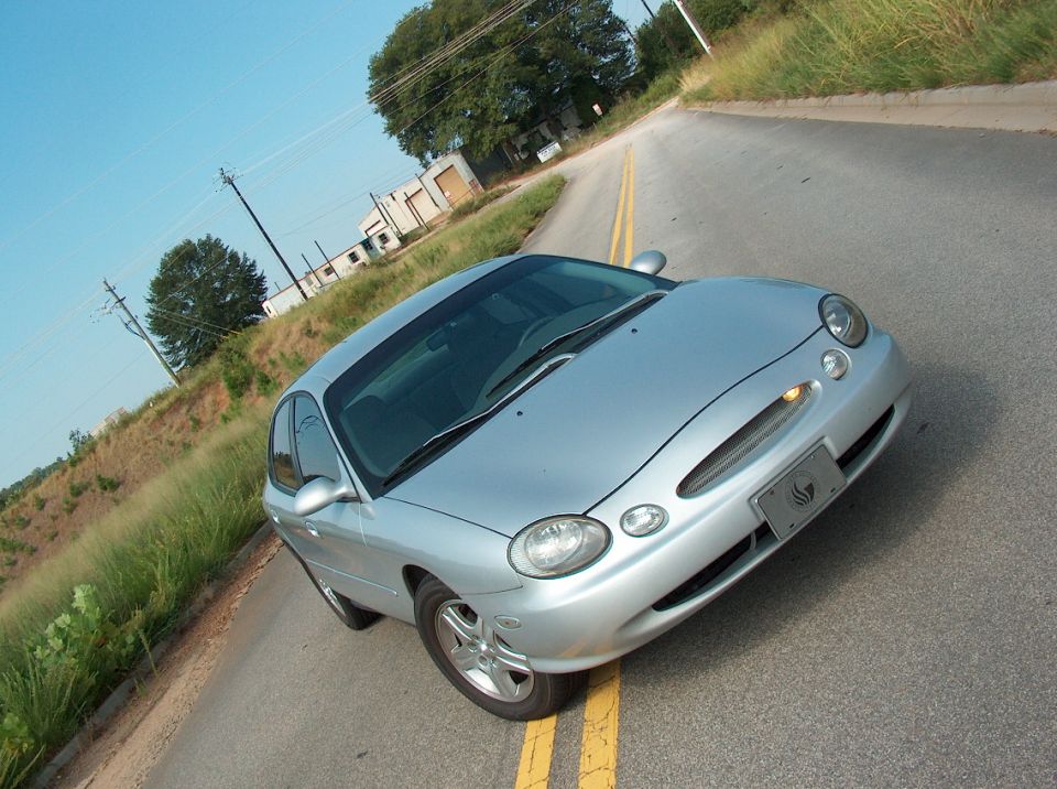 Silver sedan parked on a two-lane road with a grassy embankment and some buildings in the background