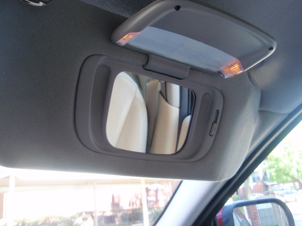 Interior view of a car's sun visor with a vanity mirror and illuminated light. Reflection shows beige seats