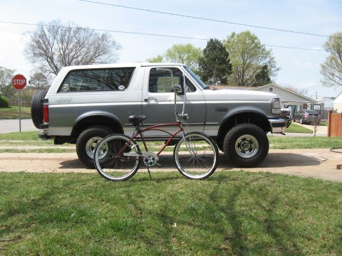1994 Ford Bronco - A two-tone Ford Bronco is parked next to a red cruiser bicycle on a sunny day.