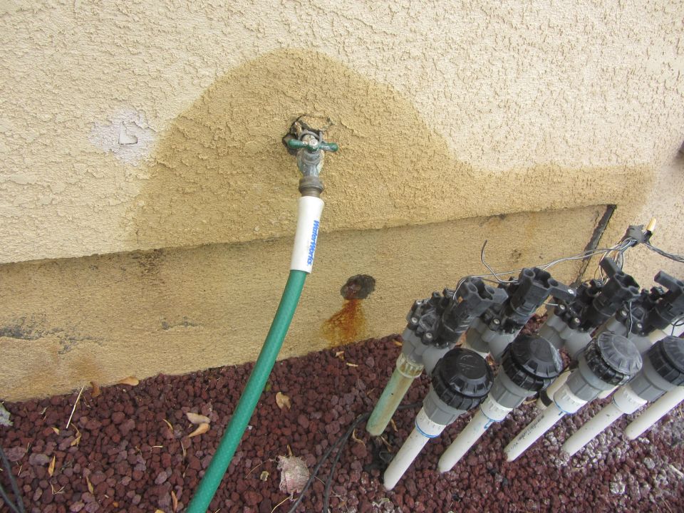 Leaky faucet with green hose attached to stucco wall, next to row of irrigation valves