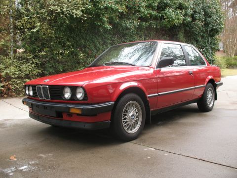 1986 BMW 3 Series - A red 1986 BMW 3 Series coupe is viewed from the front-left, parked outdoors on a paved surface with trees in the background.