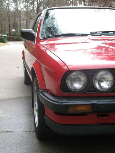 1986 BMW 3 Series - A red BMW E30 coupe is parked on a wet driveway, showing its front end and side profile.