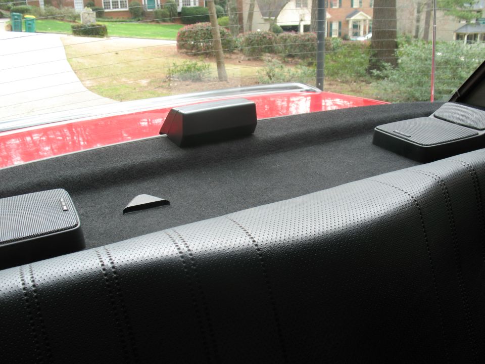 Rear parcel shelf with black carpet, two box speakers, and third brake light; red trunk lid seen through the rear window.