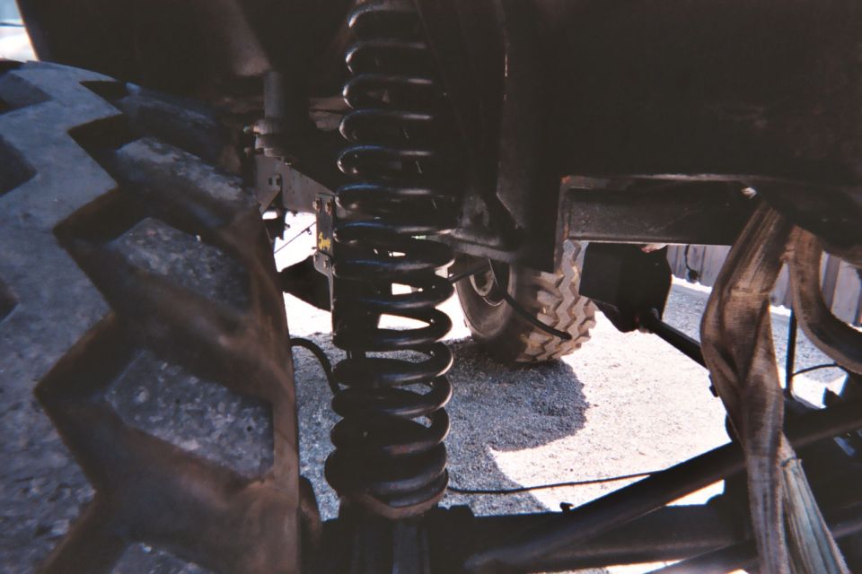 Close-up under a lifted vehicle showing black coil spring, shock, and large tire over gravel