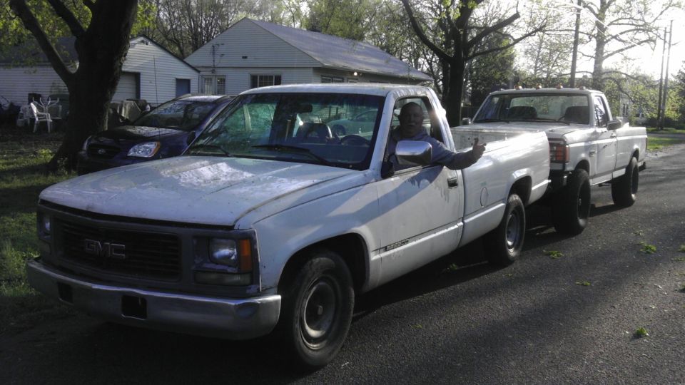 White GMC pickup truck with a man giving a thumbs up in the driver's seat, followed by another white pickup truck