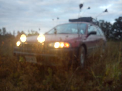 1998 Subaru Legacy - A red wagon with a roof rack and several spotlights is parked in a field as dusk settles.