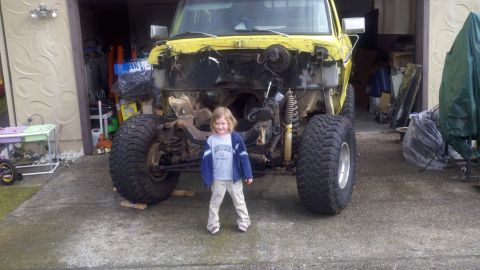 1978 Ford Bronco - A young child stands before a yellow truck undergoing significant modifications, including a removed front end and lifted suspension.