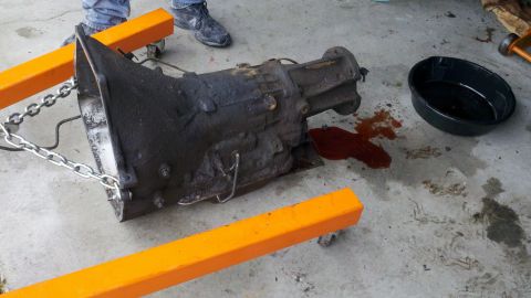 1978 Ford Bronco - A greasy transmission is being supported by an engine hoist, with a drain pan and spilled fluid nearby.