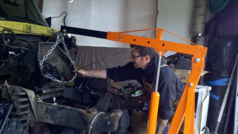 1978 Ford Bronco - A man uses an engine hoist to work on the transmission of a yellow vehicle in a garage.
