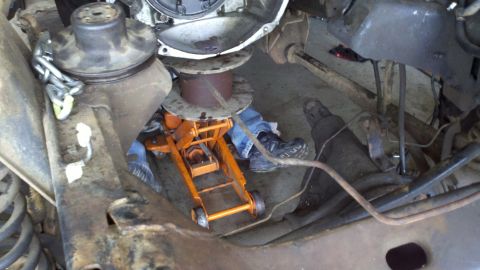 1978 Ford Bronco - A mechanic works on the transmission of a vehicle, supported by a jack and other equipment.