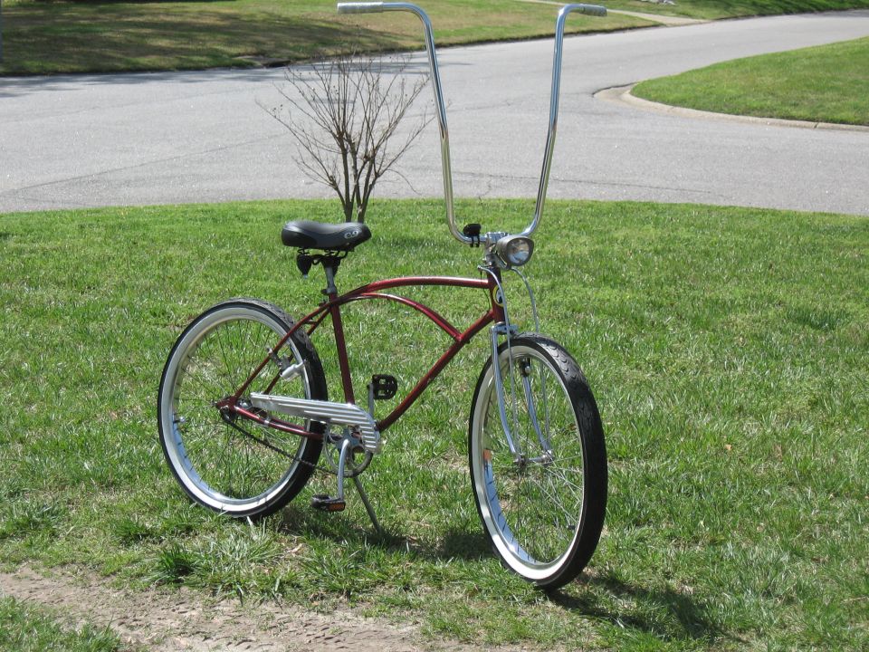Maroon cruiser bicycle with tall chrome handlebars and whitewall tires, parked on grass